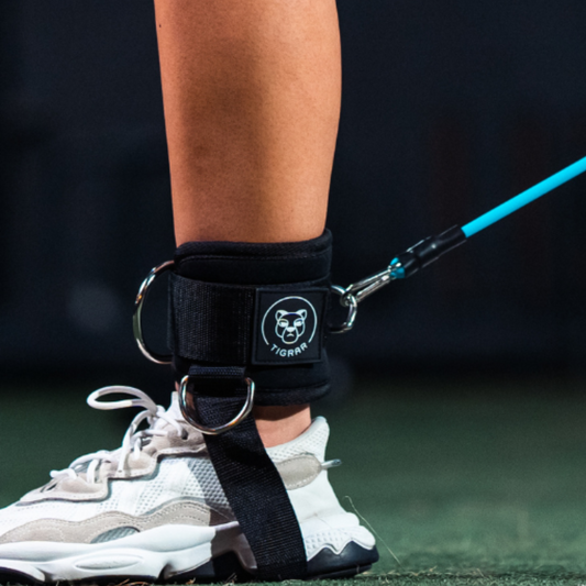 Sportieve vrouw met Tigrar enkel strap, bevestigd aan een elastische tube, in actie tijdens een gym workout.