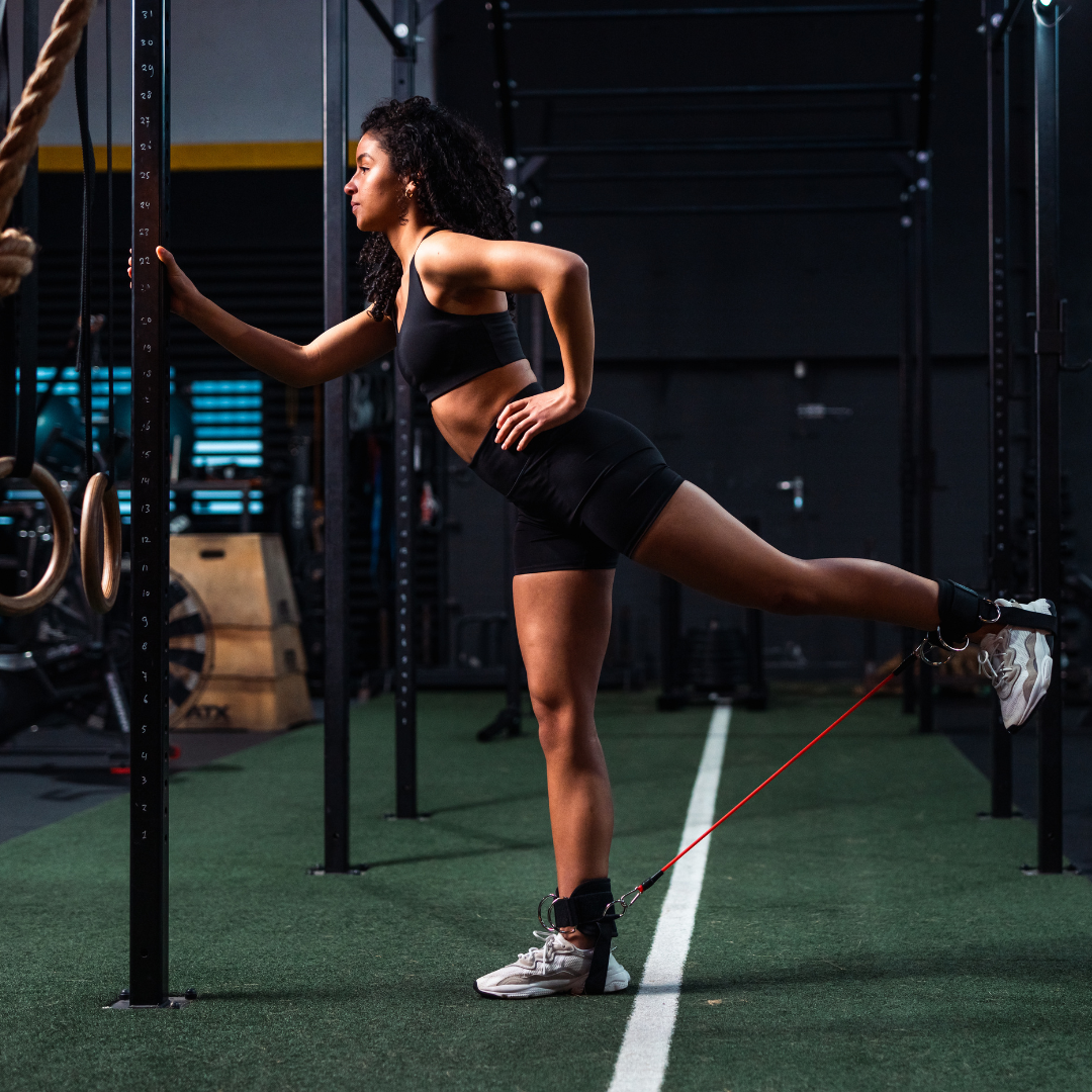 Atlete doet glute kickback in de gym, met Tigrar enkelbanden en resistance tube. Zijaanzicht laat de beenbeweging naar achter zien.