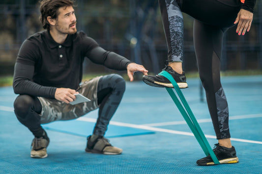 Vrouw voert standing hip flexion uit met een groene booty band om haar voeten, buiten op een  trainingsveld