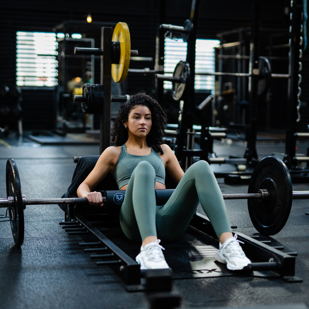 Atlete traint met een Tigrar barbell foam pad tijdens een hip thrust, fitnessapparatuur op de achtergrond.